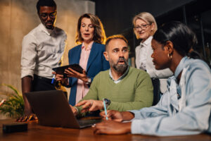 Group of five people stand around a laptop sitting on a desk